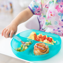 Load image into Gallery viewer, A child eats kiwi, eggs with tomatoes, and a pastry from a Bumkins Silicone Grip Dish: Blue Jelly while wearing a colorful bib.