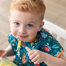 Load image into Gallery viewer, A child in a dark blue bib smiles at the camera, holding a Bumkins Spoon + Fork: Sage with spaghetti.