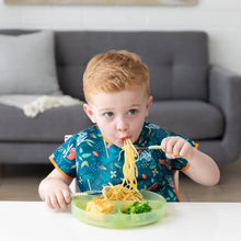 Load image into Gallery viewer, A child in a bib enjoys noodles with a fork, eating pasta and broccoli from a Bumkins Silicone Grip Dish: Green Jelly at a white table.