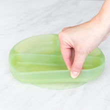 Load image into Gallery viewer, A hand holds a Bumkins Silicone Grip Dish: Green Jelly, demonstrating the suction cup bottom on a marble surface.