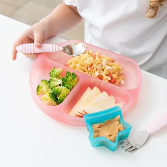 A child at a white table enjoys a meal from a Bumkins Silicone Grip Dish: Pink Jelly. A nearby blue star-shaped dish holds peanut butter.
