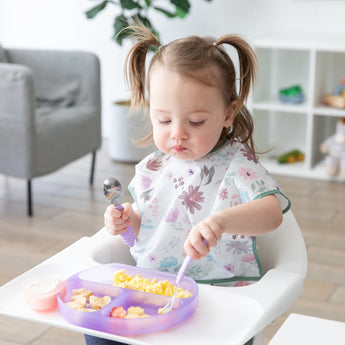 A toddler with pigtails sits in a high chair, enjoying scrambled eggs and fruit from a Bumkins Silicone Grip Dish: Purple Jelly.