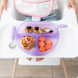 A child in a high chair has a Bumkins Silicone Grip Dish: Purple Jelly with cinnamon rolls, melon flowers, berries, and purple utensils.
