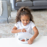 A girl in a white t-shirt removes a Bumkins Silicone Stretch Lid for Grip Dish 3 Section, Grip Plate from a white plate on a white table.