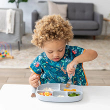 Load image into Gallery viewer, A child enjoys a meal from Bumkins Silicone Grip Dish with Lid (5 Section): Gray with a spoon, wearing a colorful bib in a cozy living room.