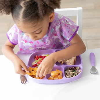 A curly-haired child enjoys snacks from a Bumkins Silicone Grip Dish with Lid (5 Section): Lavender, wearing a purple bib at a white table.