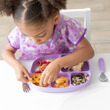 A curly-haired child enjoys snacks from a Bumkins Silicone Grip Dish with Lid (5 Section): Lavender, wearing a purple bib at a white table.