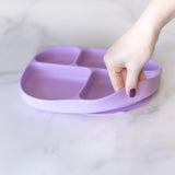 A hand holds a Bumkins Silicone Grip Dish with Lid (5 Section): Lavender, demonstrating the suction cup bottom on a marble surface.