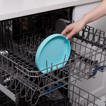 A hand places a Bumkins Silicone Grip Plate: Blue into the top rack of an open dishwasher.