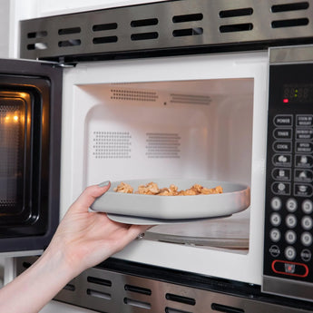 A hand places a Bumkins Silicone Grip Plate: Gray filled with food into an open microwave.