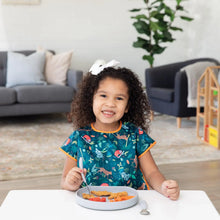 Load image into Gallery viewer, A smiling girl with curly hair enjoys chicken nuggets from a Bumkins Silicone Grip Plate: Gray at a white table in a cozy living room.
