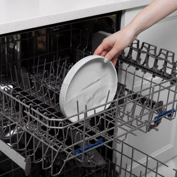 A hand places a Bumkins Silicone Grip Plate: Marble into the top rack of an open dishwasher.