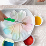 Close-up of a child painting with watercolors in cups on a Bumkins Silicone Grip Plate: Marble atop a beige patterned tablecloth.
