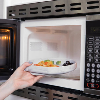 A hand places a Bumkins Silicone Grip Plate: Marble filled with food into an open microwave.