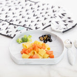 Bumkins Silicone Grip Plate: Marble with pasta, tomatoes, broccoli & olives sits in front of a bib on a marble surface with utensils nearby.
