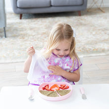 Load image into Gallery viewer, A young girl in a purple bib removes a lid from a Bumkins Silicone Grip Plate: Pink with a sandwich & strawberries on a white table.