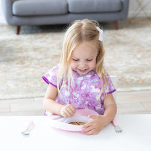 Load image into Gallery viewer, A girl in a purple bib smiles at a white table, removing a lid from a Bumkins Silicone Grip Plate: Pink while a spoon and fork rest nearby.