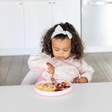 A toddler enjoys scrambled eggs and berries from a Bumkins Silicone Grip Plate: Pink while sitting in a white kitchen.
