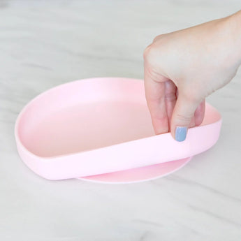 A hand holds a Bumkins Silicone Grip Plate: Pink, demonstrating the suction cup bottom on a marble surface.