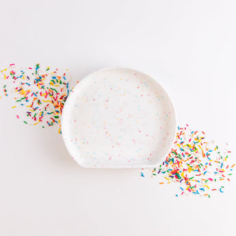 Bumkins Silicone Grip Plate: Vanilla Sprinkle lays on top of a pile of sprinkles in a diagonal line on a white background.