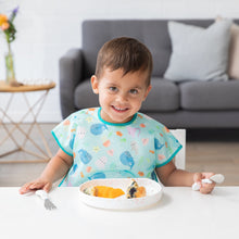 Load image into Gallery viewer, A smiling child sits at a white table wearing a colorful bib, ready to eat from a Bumkins Silicone Grip Plate: Vanilla Sprinkle.