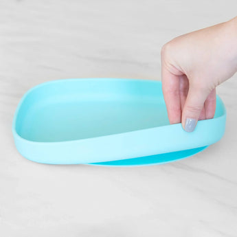 A hand holds a Bumkins Silicone Grip Tray: Blue, demonstrating the suction cup bottom on a marble surface.