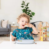 A curly-haired child enjoys a sandwich from Bumkins Silicone Grip Tray: Gray, wearing a colorful bib with a plant and wood toy house nearby.