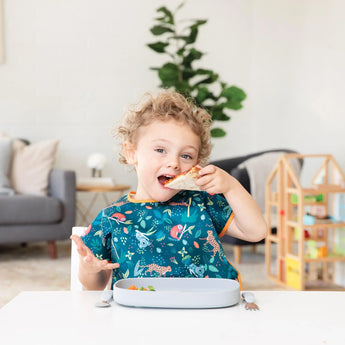 A curly-haired child enjoys a sandwich from Bumkins Silicone Grip Tray: Gray, wearing a colorful bib with a plant and wood toy house nearby.