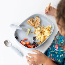 Load image into Gallery viewer, A child in a colorful bib enjoys eggs, fruit, and oats from a Bumkins Silicone Grip Tray: Gray at a white table with utensils nearby.