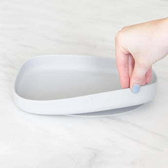 A hand holds a Bumkins Silicone Grip Tray: Gray, demonstrating the suction cup bottom on a marble surface.
