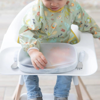 A child in a high chair wearing a green patterned bib reaches for a lid covering a Bumkins Silicone Grip Tray: Marble with utensils nearby.