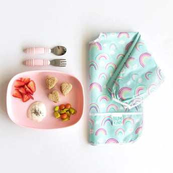 Bumkins Silicone Grip Tray: Pink with fruit and yogurt sits next to pink utensils and a folded rainbow-print bib on a white background.