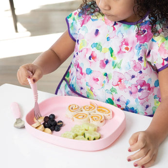 A child with a colorful bib sits at a white table, eating rolled sandwiches and fruit from a Bumkins Silicone Grip Tray: Pink with a fork.