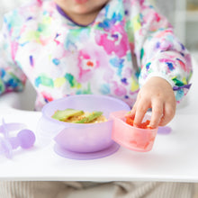 Load image into Gallery viewer, A child in a bib reaches for food from a Bumkins Silicone Little Dipper Combo 2 Pack: Magenta Jelly attached to a bowl on a high chair tray.