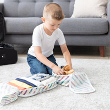 Load image into Gallery viewer, A child organizes clothes into Bumkins Packing Cube 2 Pack: Mickey & Friends Varsity on a carpeted floor with nearby luggage and a gray couch.