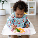 A toddler in a bib sits in a high chair, eating from Bumkins Silicone Plate + Spoon Set: Disney Princess with veggies, nuggets, and potatoes.