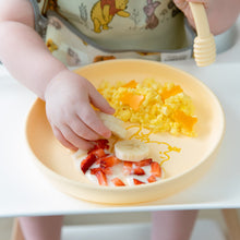Load image into Gallery viewer, A child wearing a bib in a high chair enjoys breakfast from a Bumkins Silicone Plate + Spoon Set: Disney Winnie the Pooh.