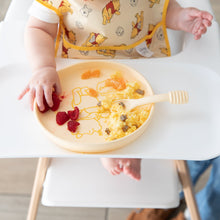 Load image into Gallery viewer, A baby in a high chair enjoys eggs and raspberries from a Bumkins Silicone Plate + Spoon Set: Disney Winnie the Pooh while wearing a bib.