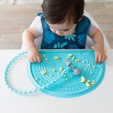 Load image into Gallery viewer, A boy wearing a Bumkins Starter Bib 2 Pack: Dinosaurs & Blue Tropic plays with scrambled eggs on a blue silicone placemat.