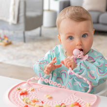 Load image into Gallery viewer, A bright-eyed baby chews a pink toddler fork attached to a Bumkins Silicone Accessory Tether 2-Pack: Pink on a pink placemat with fruit.