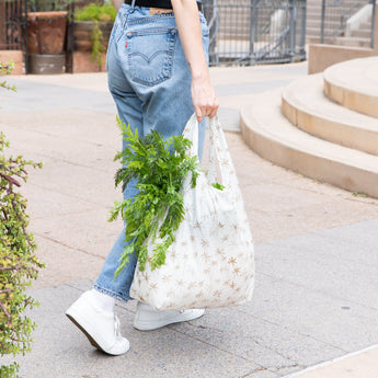 A person strolls outside on a paved path wearing jeans and white sneakers, carrying a Bumkins Packable Tote Bag: Palm Daze filled with greens.