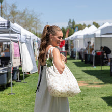 Load image into Gallery viewer, A woman in a white dress with a ponytail carries the Bumkins Packable Tote Bag: Palm Daze at a sunny outdoor market.