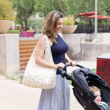 Load image into Gallery viewer, A smiling woman pushes a stroller with a Bumkins Packable Tote Bag: Palm Daze on her shoulder against urban greenery.
