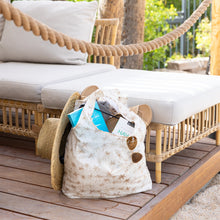 Load image into Gallery viewer, A Bumkins Packable Tote Bag: Palm Daze filled with sunglasses, books, and a straw hat sits by a wicker outdoor sofa on a wooden deck.