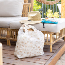 Load image into Gallery viewer, A woven straw hat and books on a wicker sofa sit near Bumkins Packable Tote Bag: Palm Daze on a wooden deck and greenery in the background.