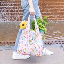 Load image into Gallery viewer, A person in jeans and white sneakers carries Bumkins Packable Tote Bag: Boots filled with flowers and vegetables, strolling past a brick wall.