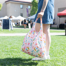 Load image into Gallery viewer, A person carries a vibrant Bumkins Packable Tote Bag: Boots in a lively park, with tents and people in the background.