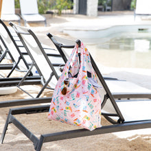 Load image into Gallery viewer, A Bumkins Packable Tote Bag: Boots hangs on a pool lounge chair. Sunglasses dangle from its side, with empty chairs behind.