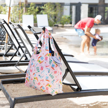 Load image into Gallery viewer, A Bumkins Packable Tote Bag: Boots hangs on a lounge chair by the pool while someone holds a child near the water.