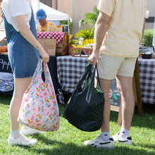 Load image into Gallery viewer, Two people holding bags, one a Bumkins Packable Tote Bag: Boots, at an outdoor market, with a stall and products displayed behind them.